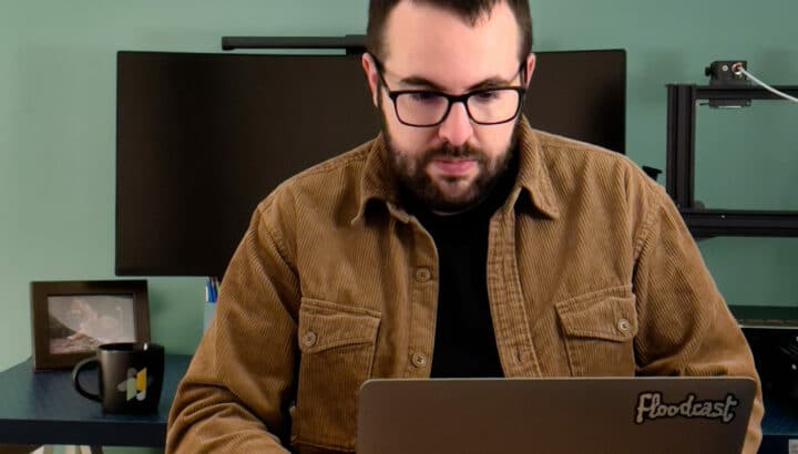 Homme avec lunettes et veste marron devant un ordinateur portable, imprimante 3D en arrière-plan