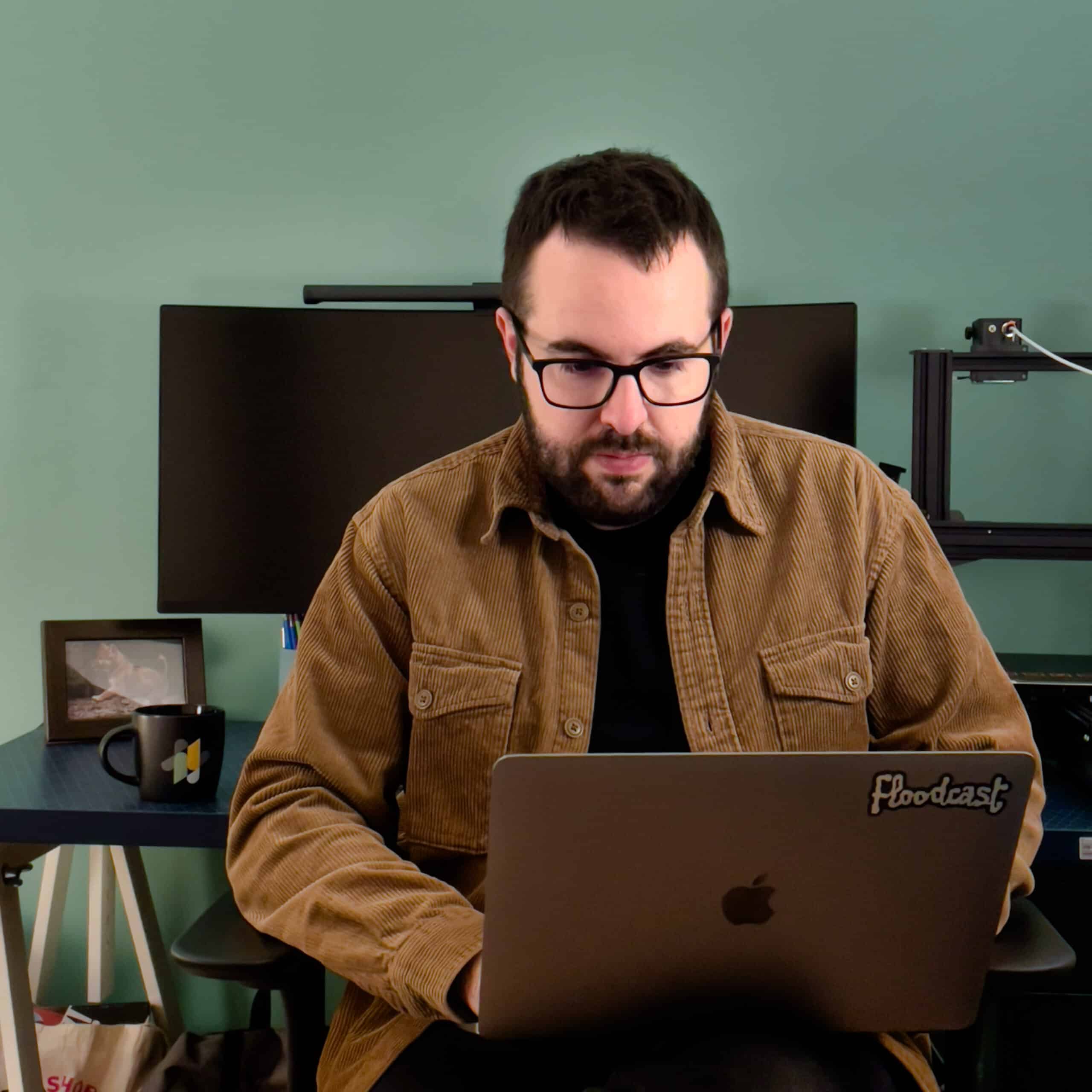 Homme avec lunettes et veste marron devant un ordinateur portable, imprimante 3D en arrière-plan