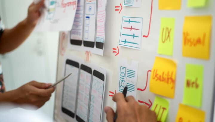 Mains disposant des post-it colorés (UX, Usability, Visual) autour de croquis de wireframes mobiles