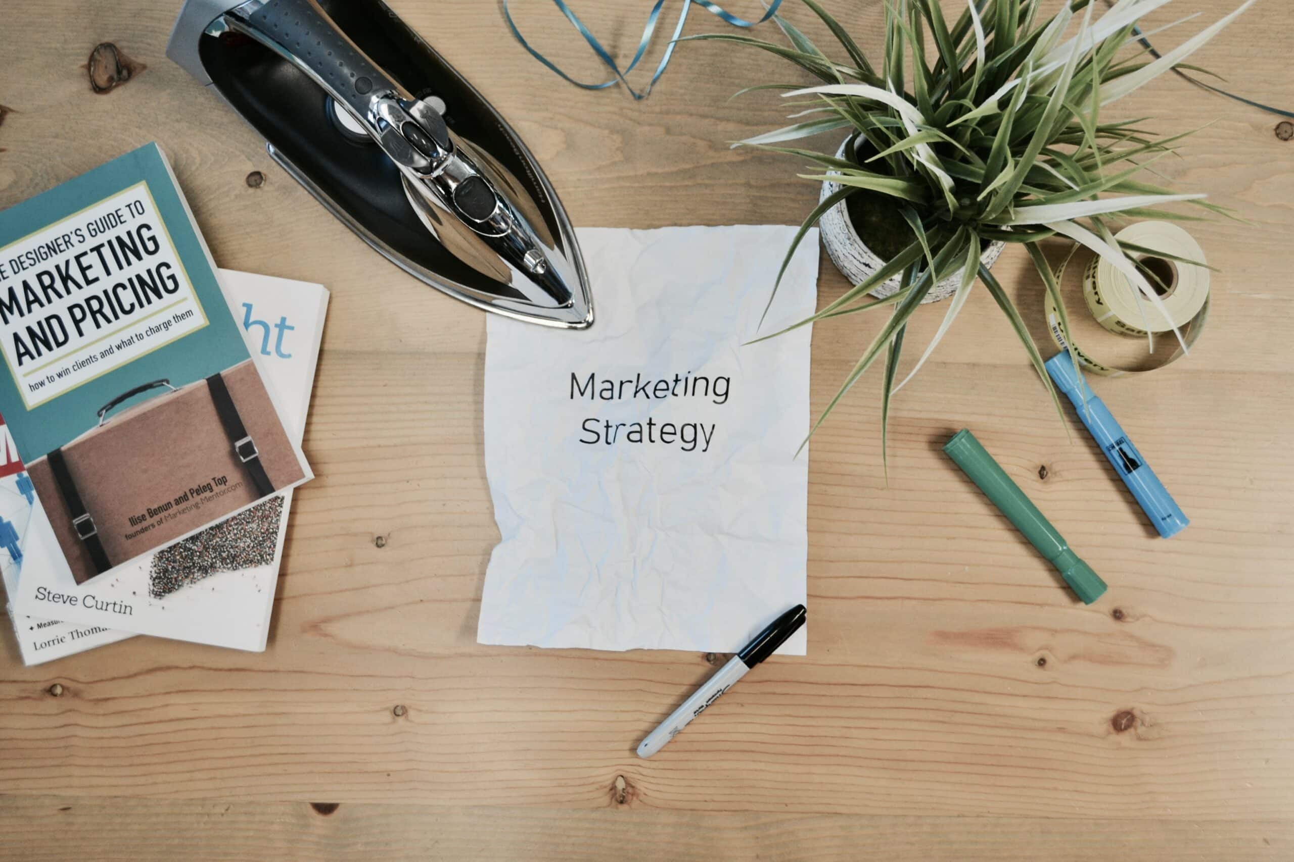 Papier froissé "Marketing Strategy" entouré d'un livre de design et d'une plante, sur bureau en bois