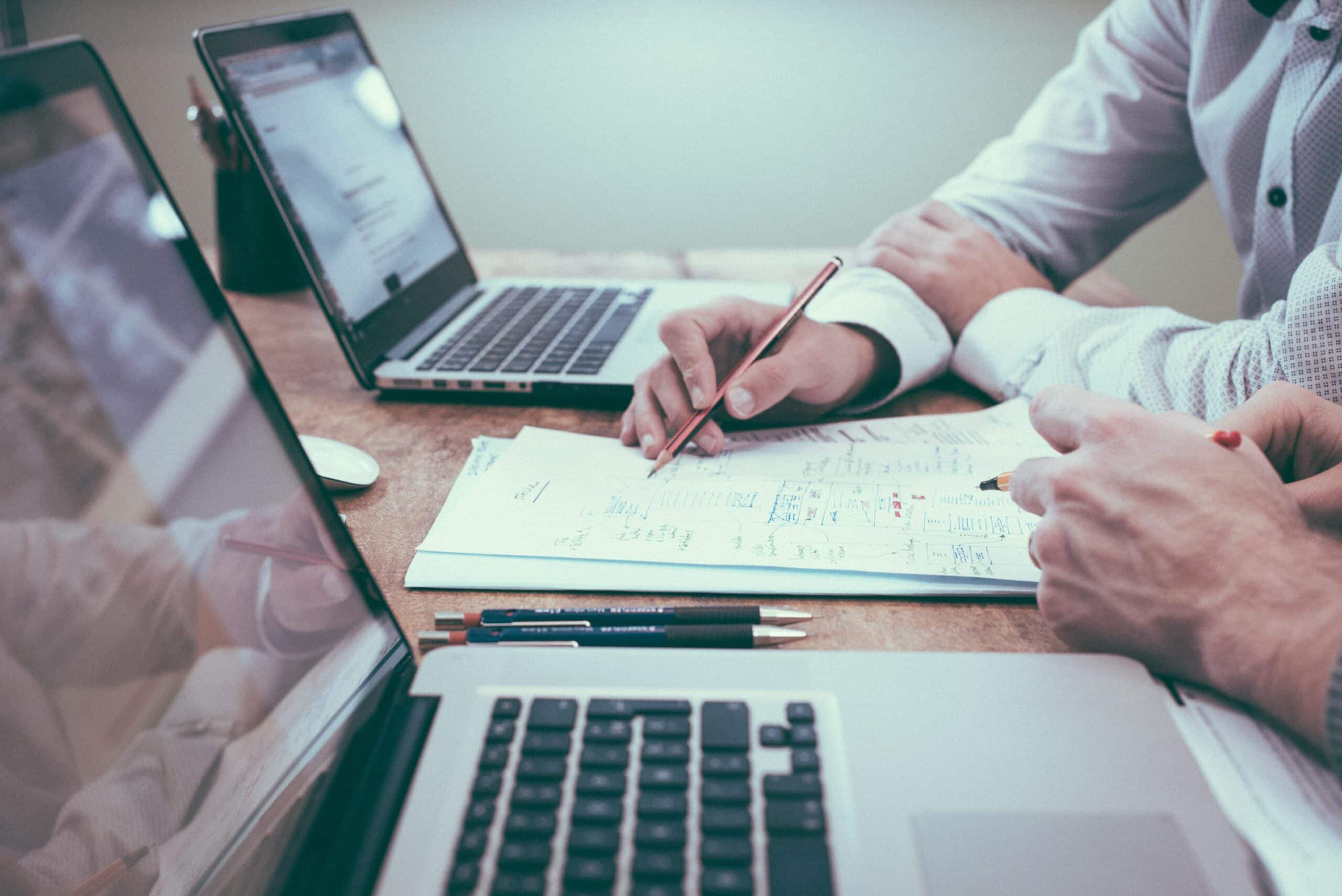 Mains annotant un cahier de planification entre deux ordinateurs portables