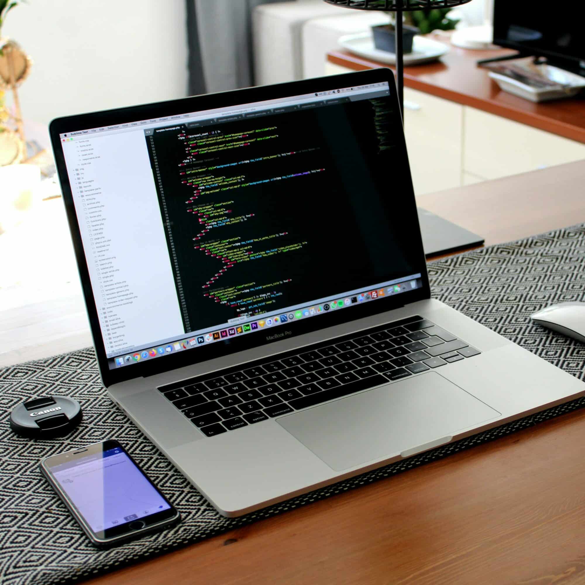 MacBook Pro affichant du code, bouchon d'objectif Canon et smartphone à côté