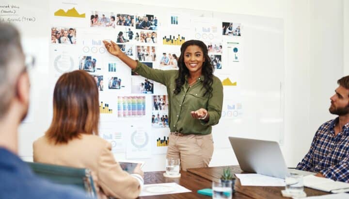 Femme souriante montrant des graphiques affichés au mur devant des collègues assis