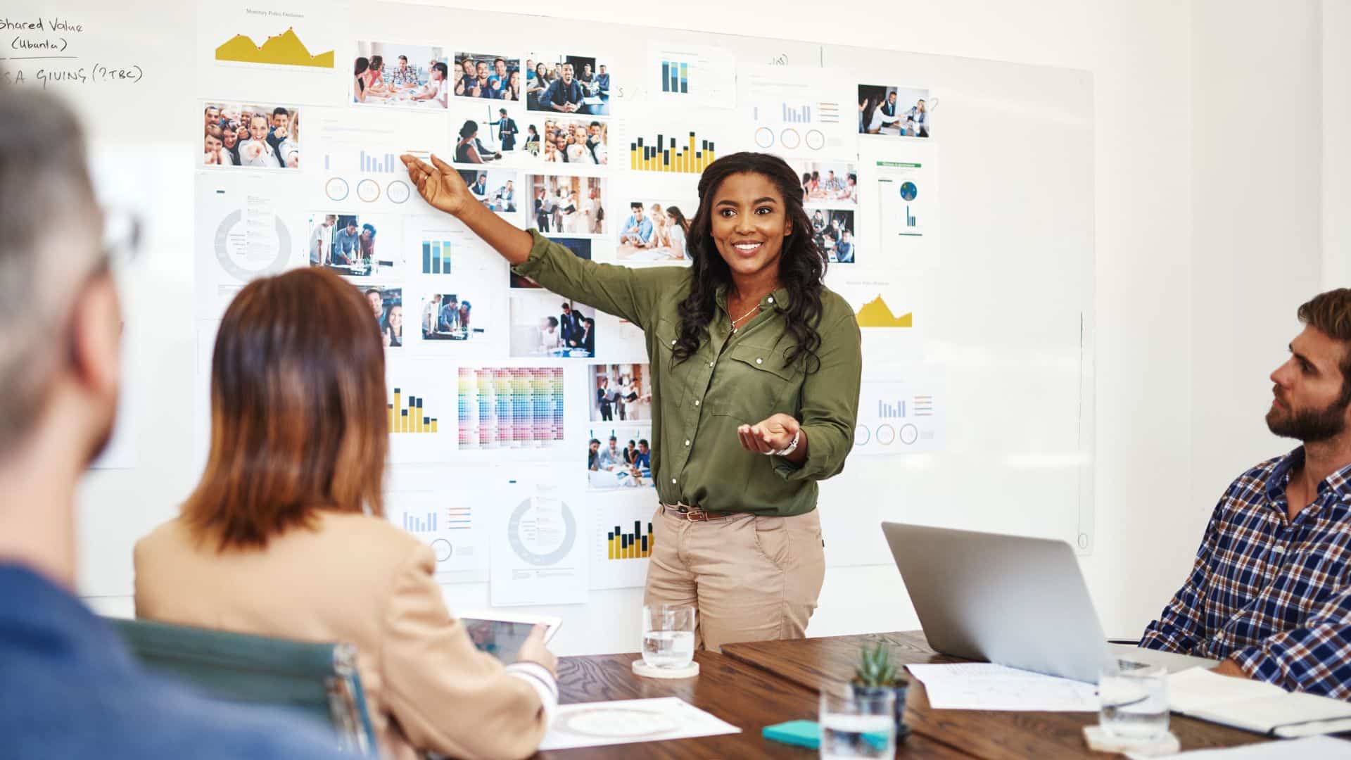 Femme souriante montrant des graphiques affichés au mur devant des collègues assis