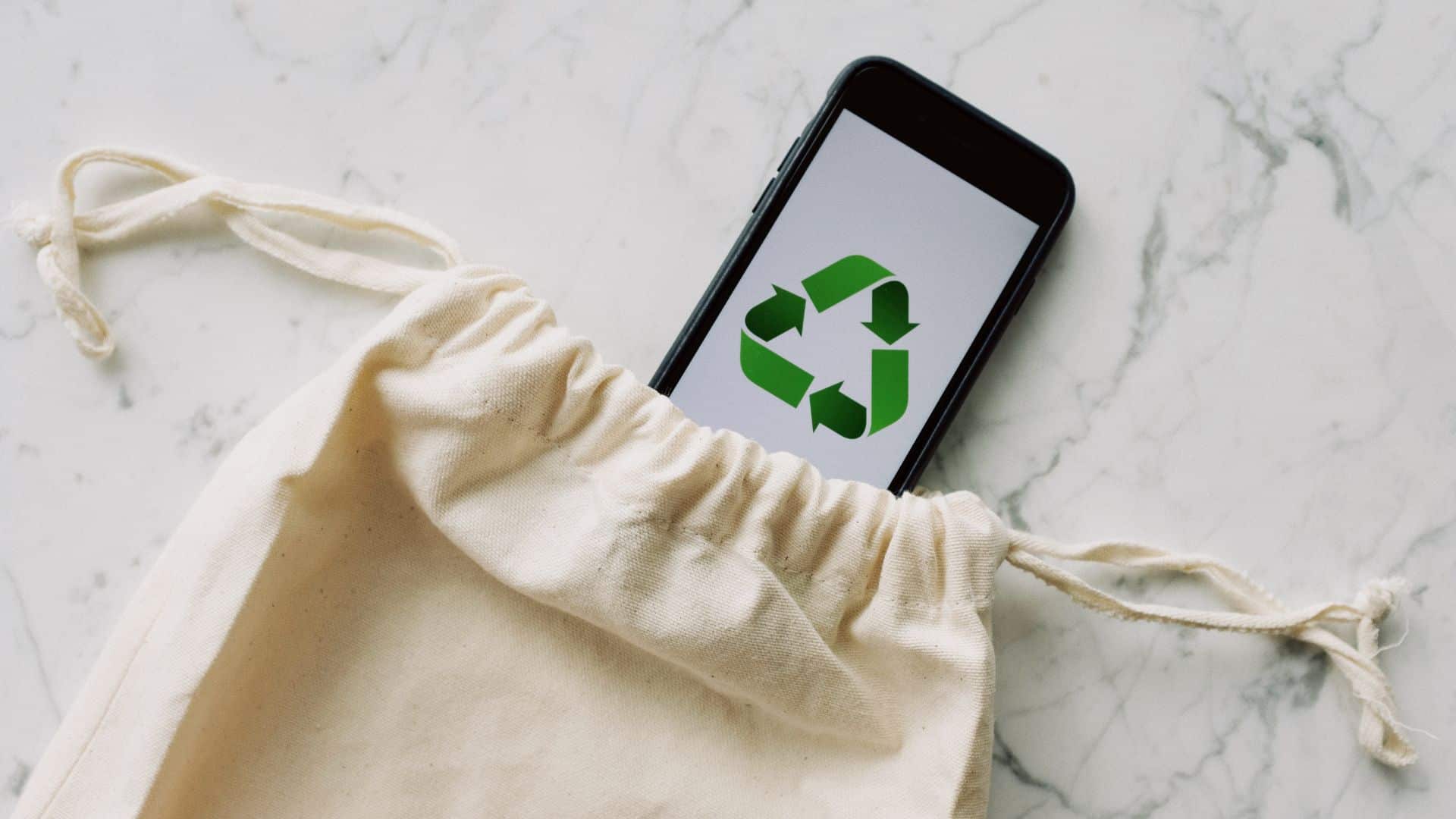 Smartphone affichant un symbole de recyclage vert, posé dans un sac en tissu sur fond marbré