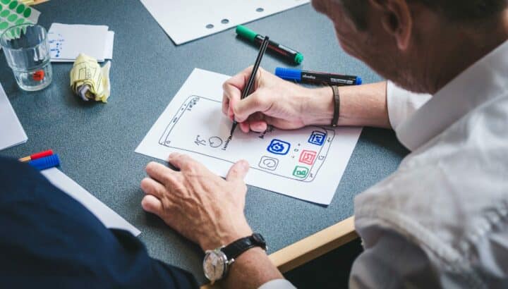 Homme esquissant une interface d'application mobile sur papier, marqueurs colorés sur le bureau