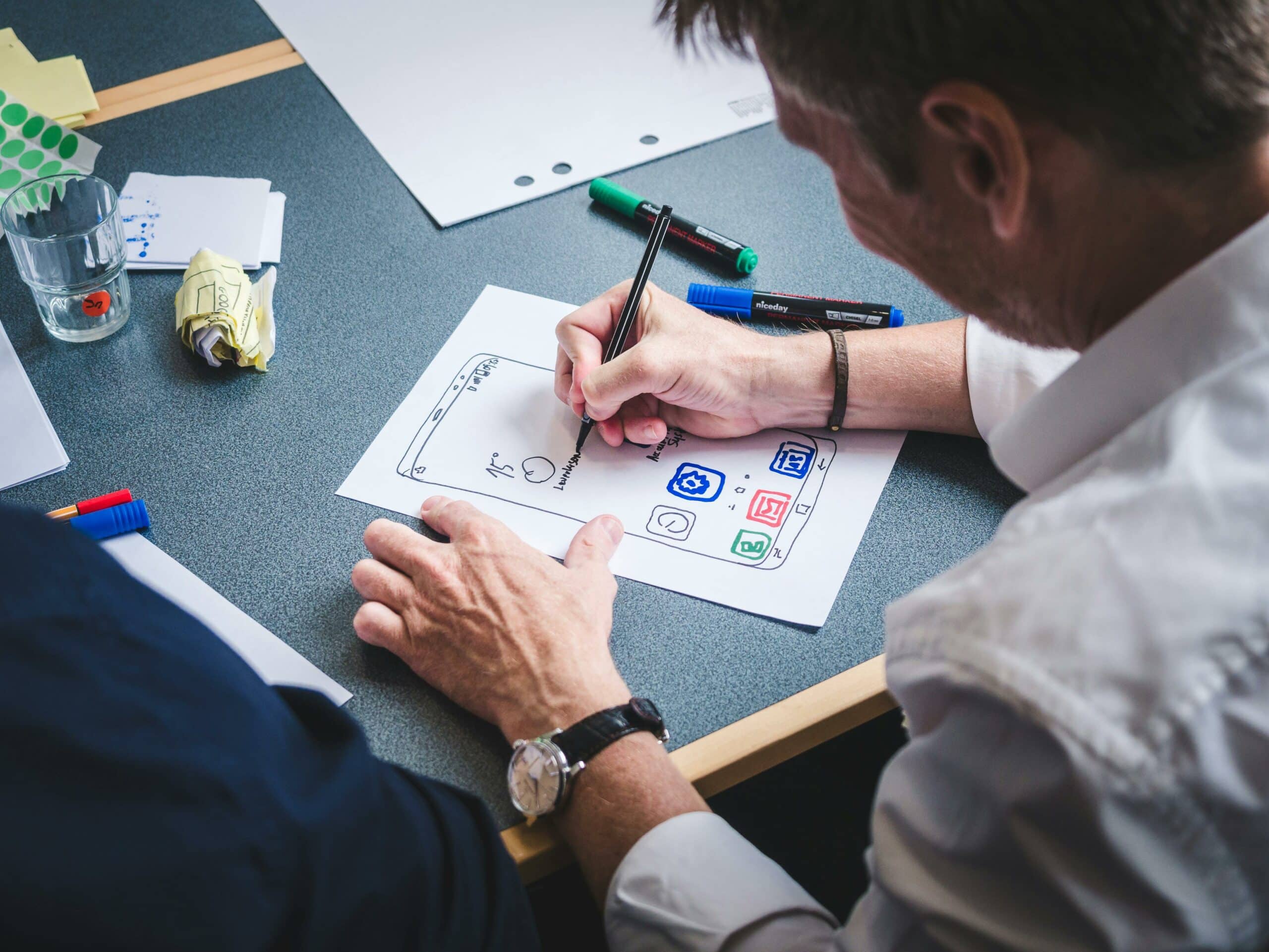 Homme esquissant une interface d'application mobile sur papier, marqueurs colorés sur le bureau