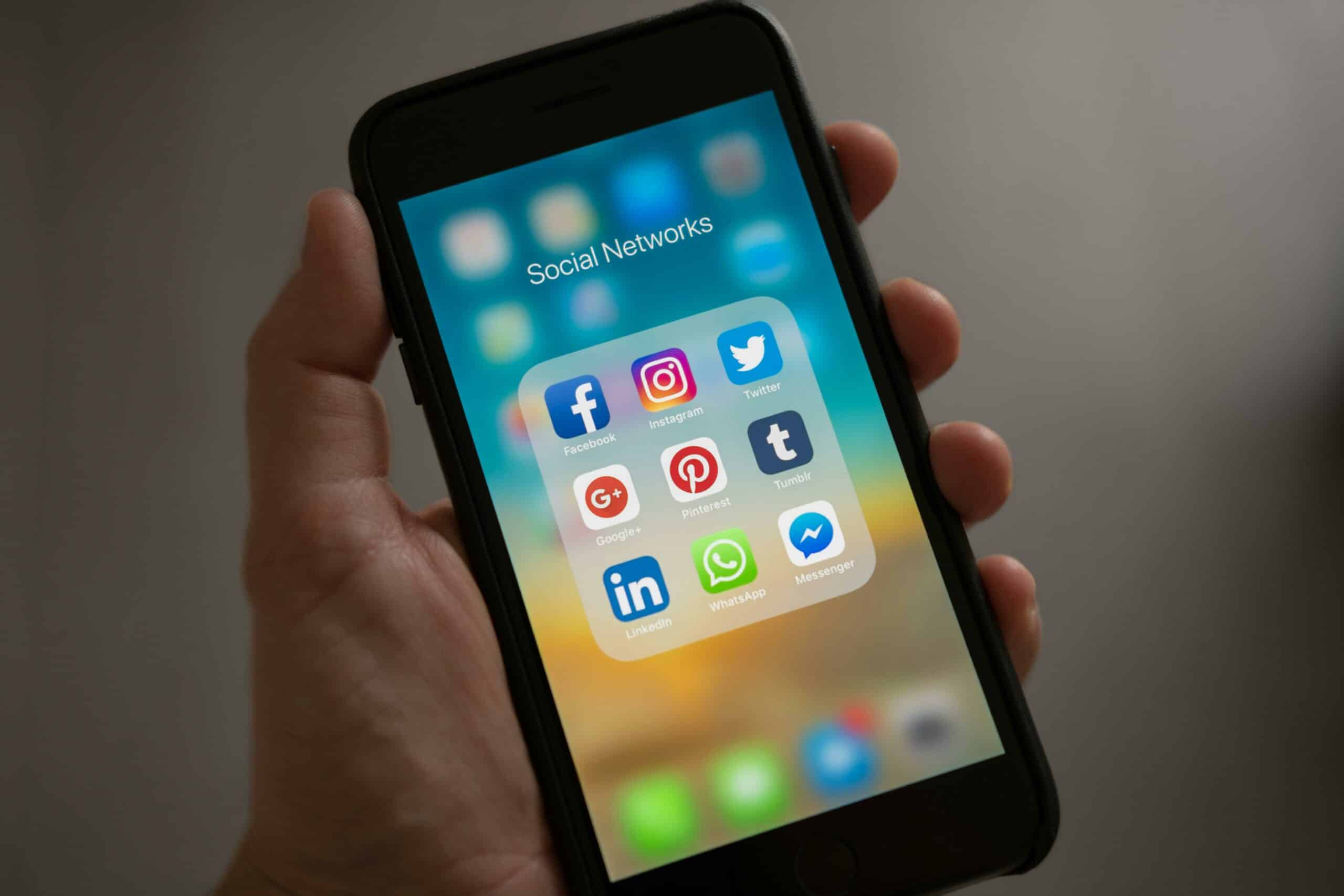 Main tenant un smartphone avec un dossier d'applications intitulé "Social Networks" (Facebook, Instagram, Twitter, etc.)