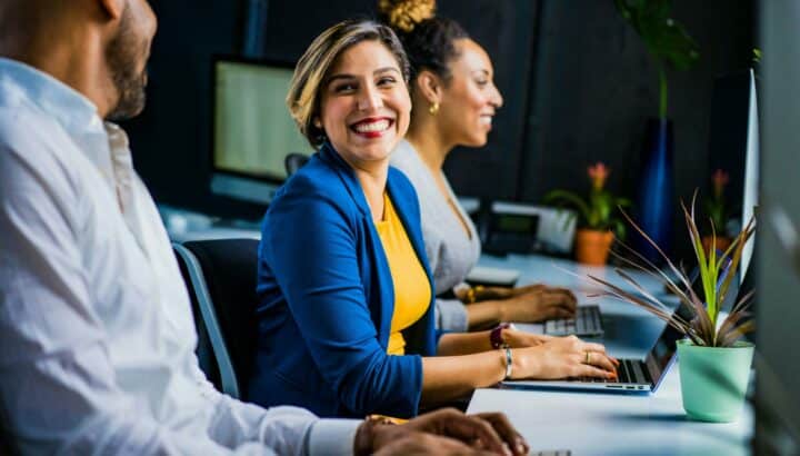 Trois collègues au bureau devant des ordinateurs, femme souriante au centre