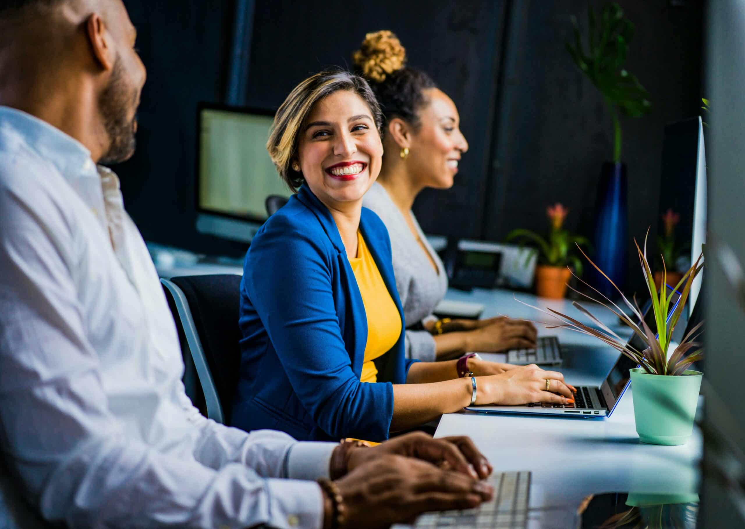 Trois collègues au bureau devant des ordinateurs, femme souriante au centre