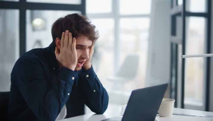 Homme en chemise bleu marine se tenant la tête, l'air stressé, devant un ordinateur portable dans un bureau lumineux