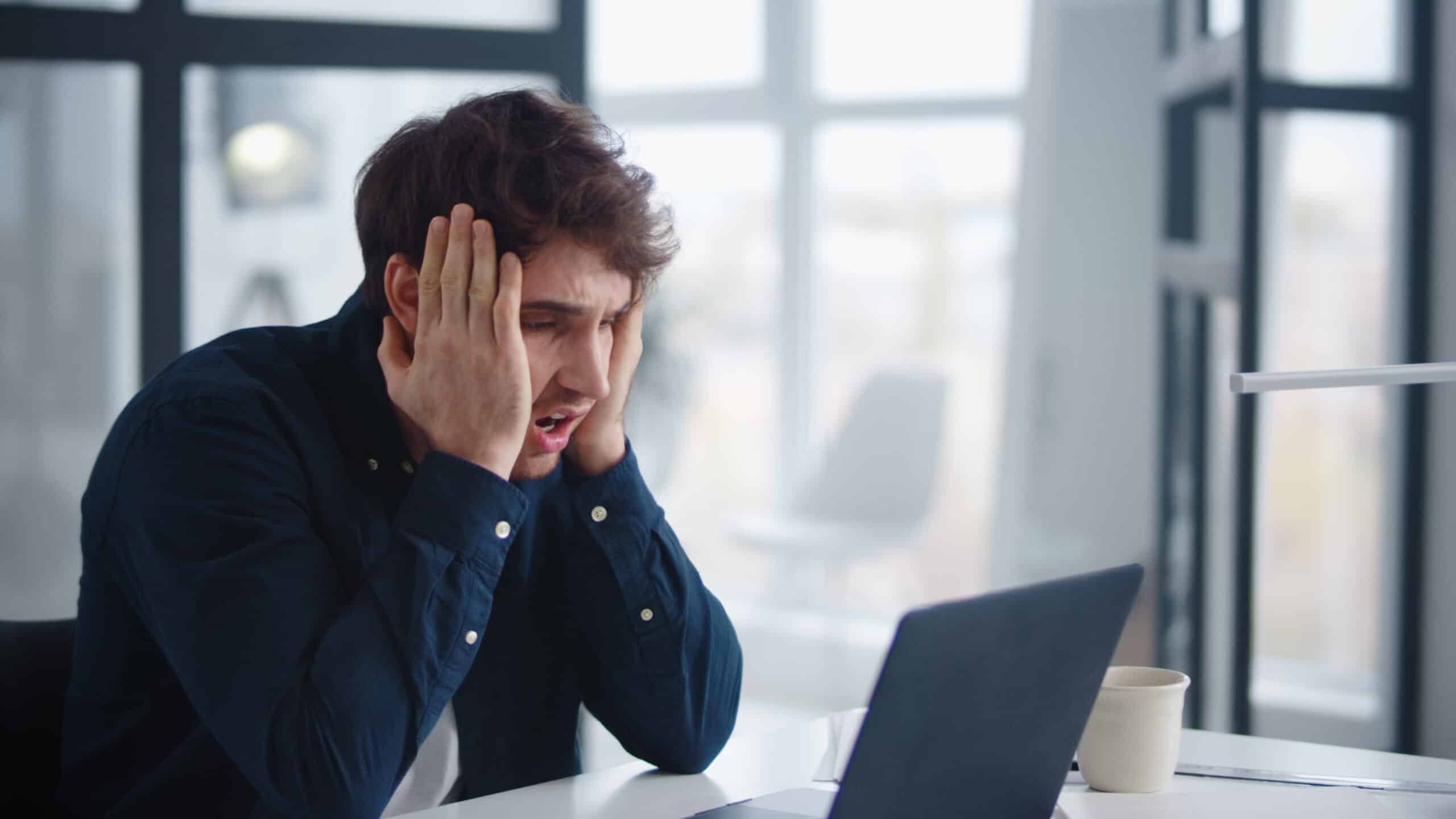 Homme en chemise bleu marine se tenant la tête, l'air stressé, devant un ordinateur portable dans un bureau lumineux