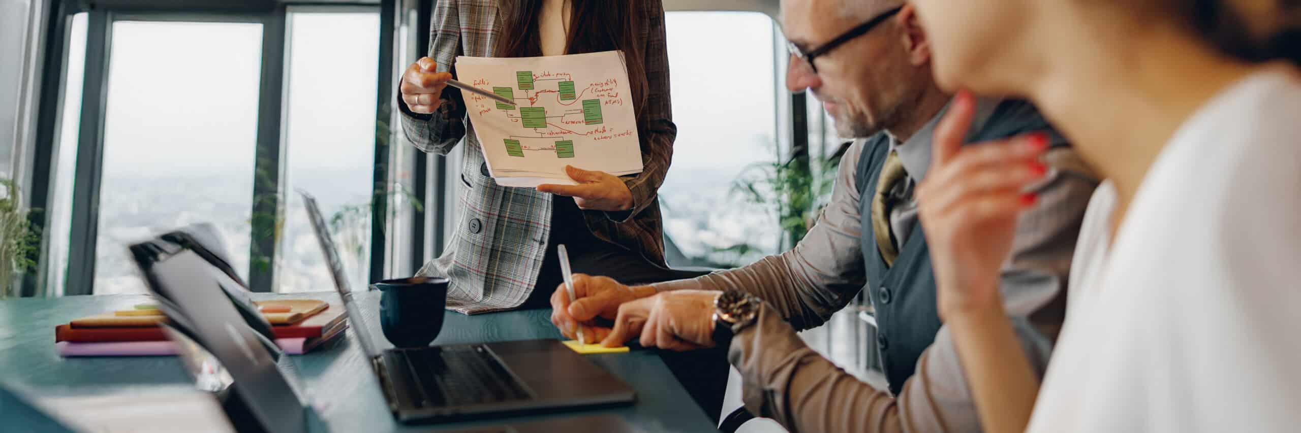 Femme tenant une feuille avec un schéma de processus annoté, deux collègues assis à une table avec ordinateurs portables, grande baie vitrée en arrière-plan