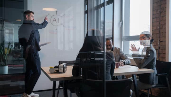 Homme dessinant un schéma "AI" sur un tableau blanc dans un bureau vitré, deux collègues discutant à une table en arrière-plan avec un casque de réalité virtuelle