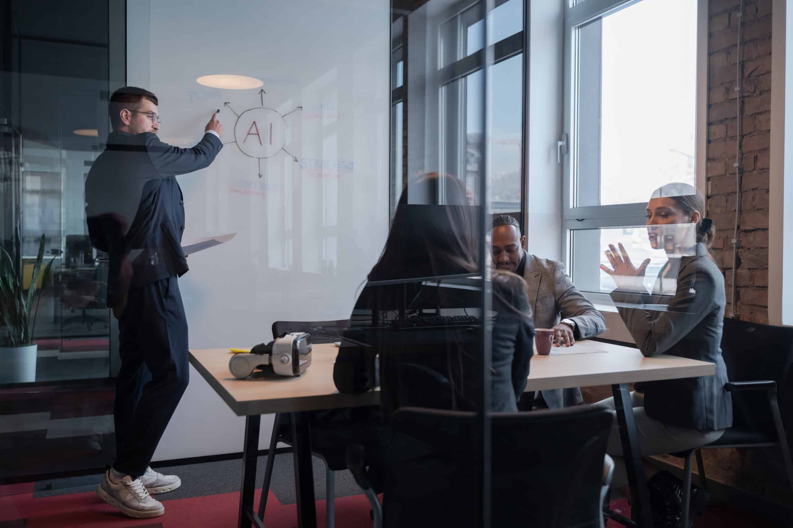Homme dessinant un schéma "AI" sur un tableau blanc dans un bureau vitré, deux collègues discutant à une table en arrière-plan avec un casque de réalité virtuelle