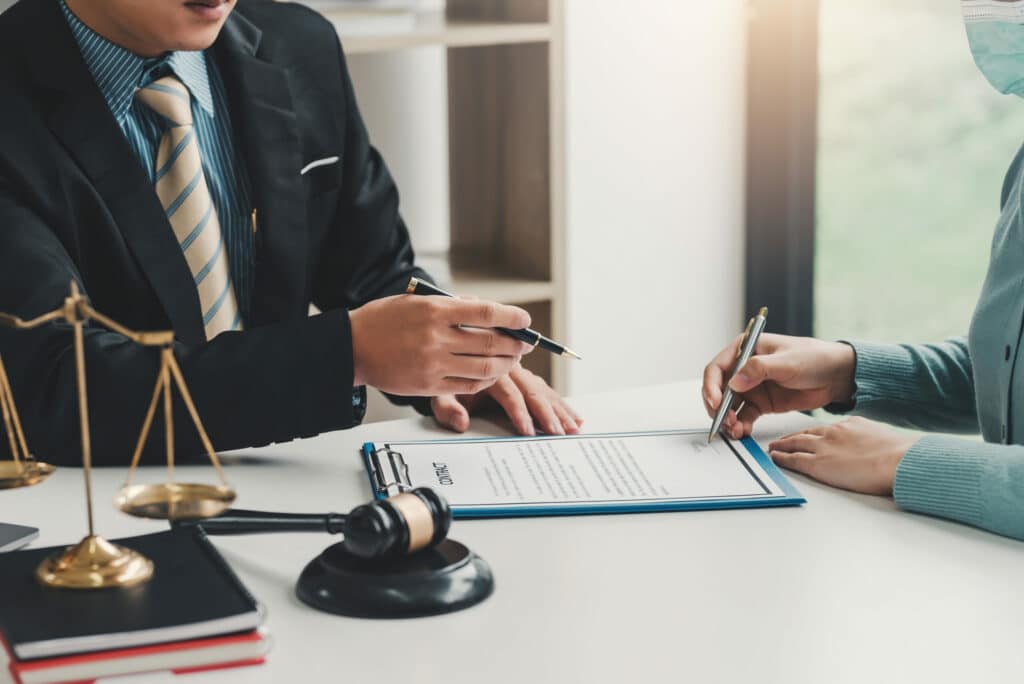 Deux personnes signant un contrat sur presse-papier bleu, avocat en costume tenant un stylo, balance de la justice et marteau de juge au premier plan