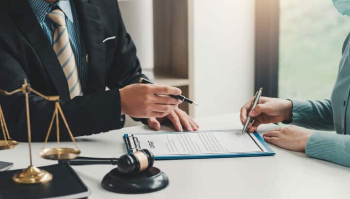 Deux personnes signant un contrat sur presse-papier bleu, avocat en costume tenant un stylo, balance de la justice et marteau de juge au premier plan