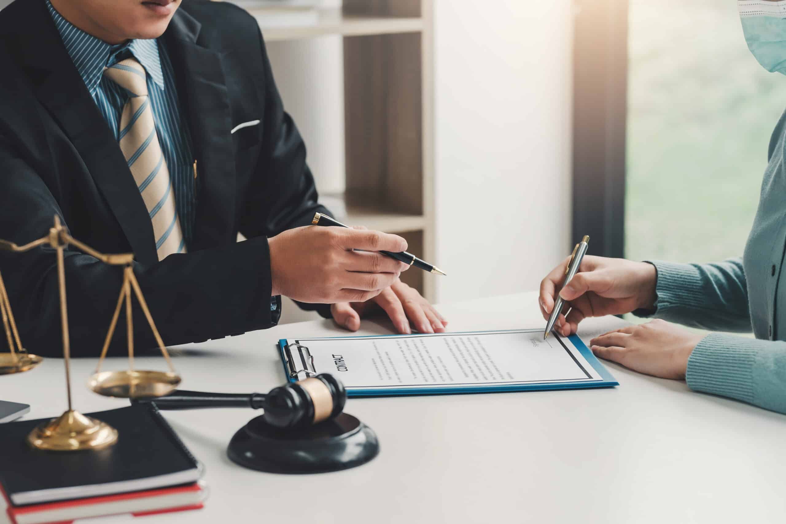 Deux personnes signant un contrat sur presse-papier bleu, avocat en costume tenant un stylo, balance de la justice et marteau de juge au premier plan