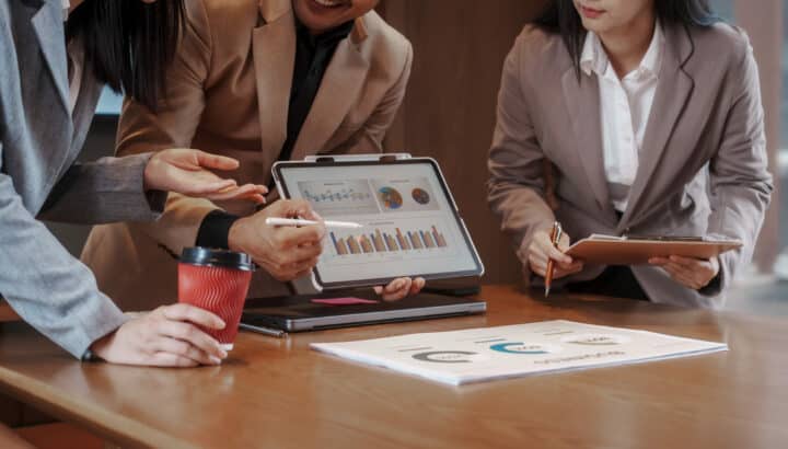 Trois personnes en tenue professionnelle penchées sur une tablette affichant des graphiques et diagrammes, café et documents sur la table