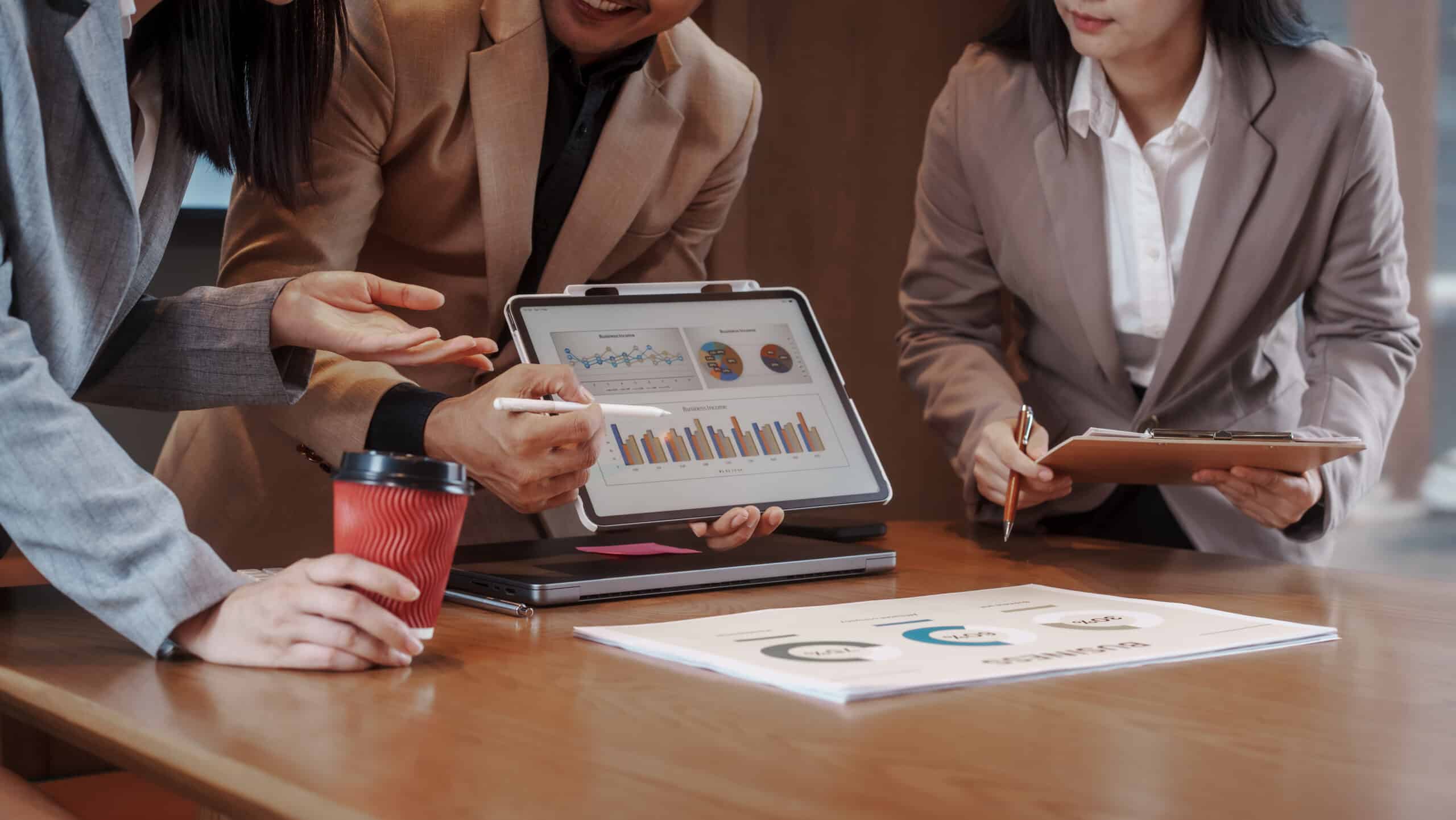 Trois personnes en tenue professionnelle penchées sur une tablette affichant des graphiques et diagrammes, café et documents sur la table