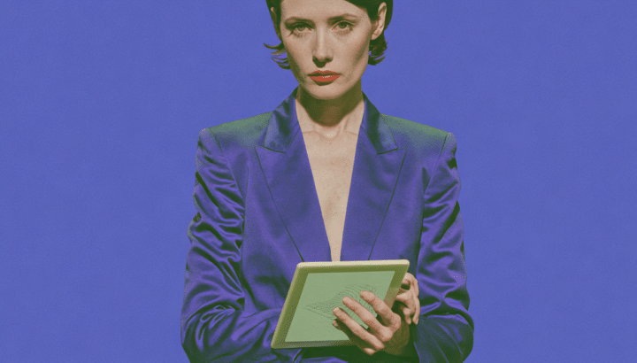 Femme en tailleur violet tenant une tablette, debout devant un bureau et une chaise, fond bleu-violet