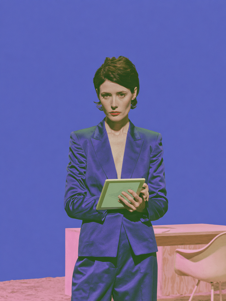 Femme en tailleur violet tenant une tablette, debout devant un bureau et une chaise, fond bleu-violet