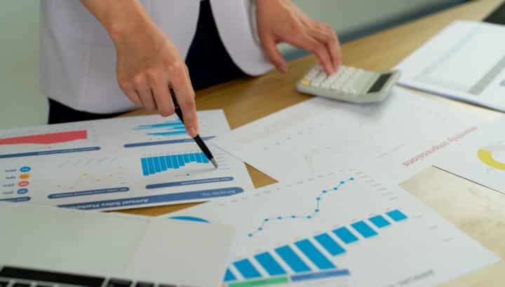 Main tenant un stylo pointant vers des graphiques de performance imprimés, calculatrice et rapports colorés étalés sur un bureau