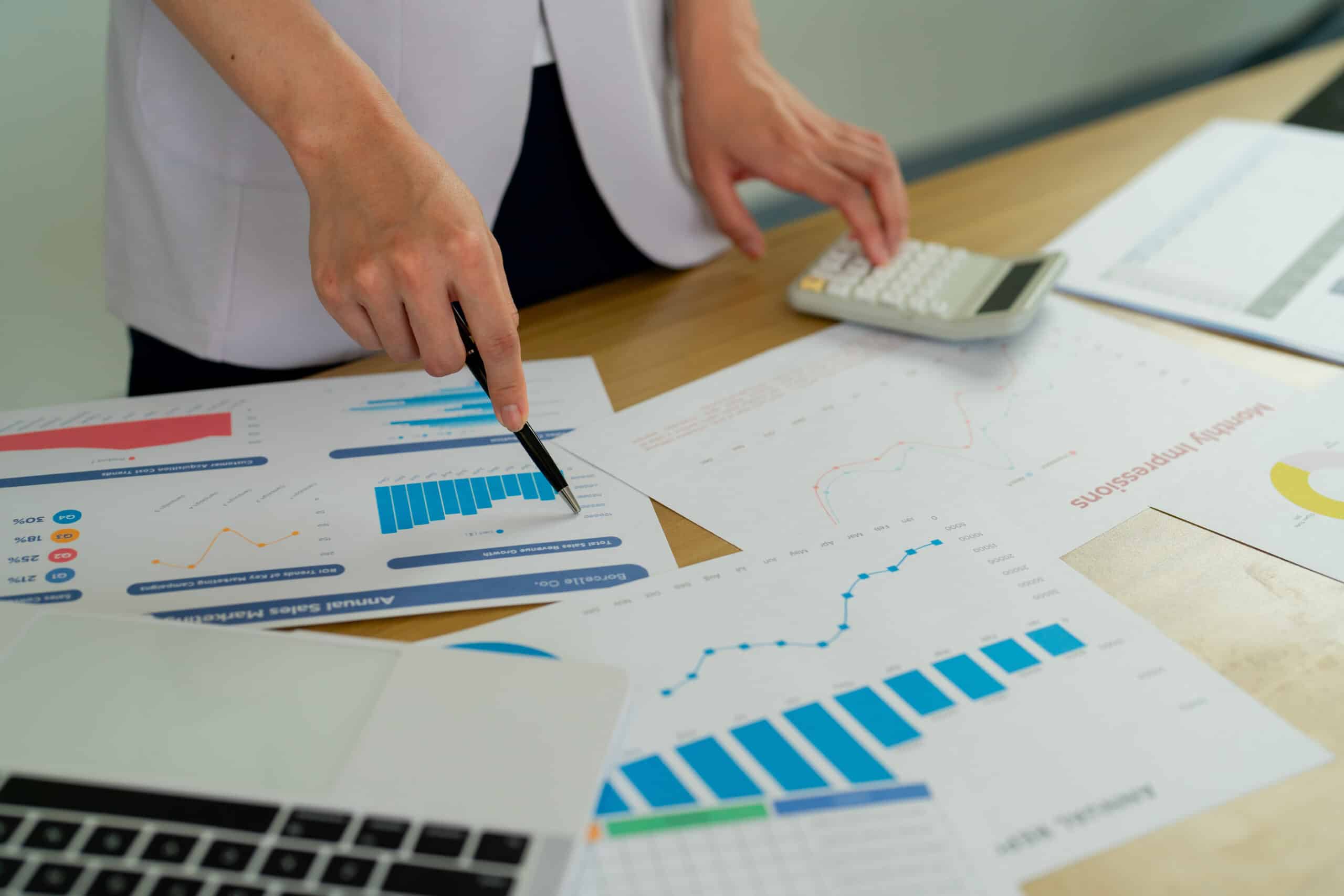 Main tenant un stylo pointant vers des graphiques de performance imprimés, calculatrice et rapports colorés étalés sur un bureau