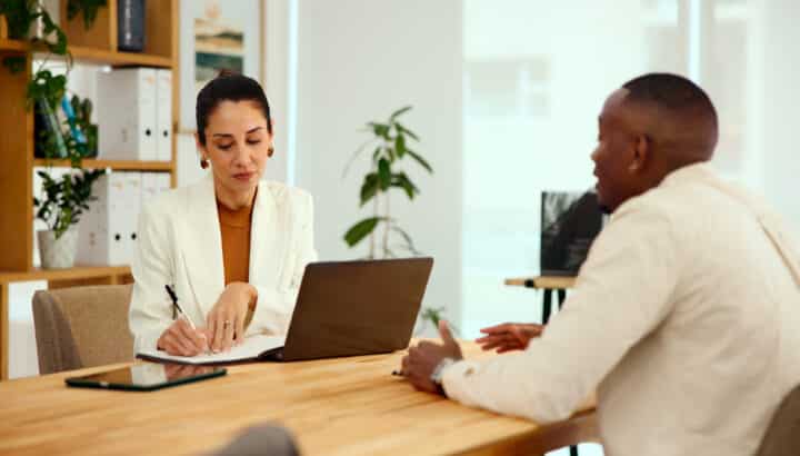 Femme en blazer blanc écrivant sur un carnet à côté d'un ordinateur portable, en réunion avec un homme, bureau lumineux