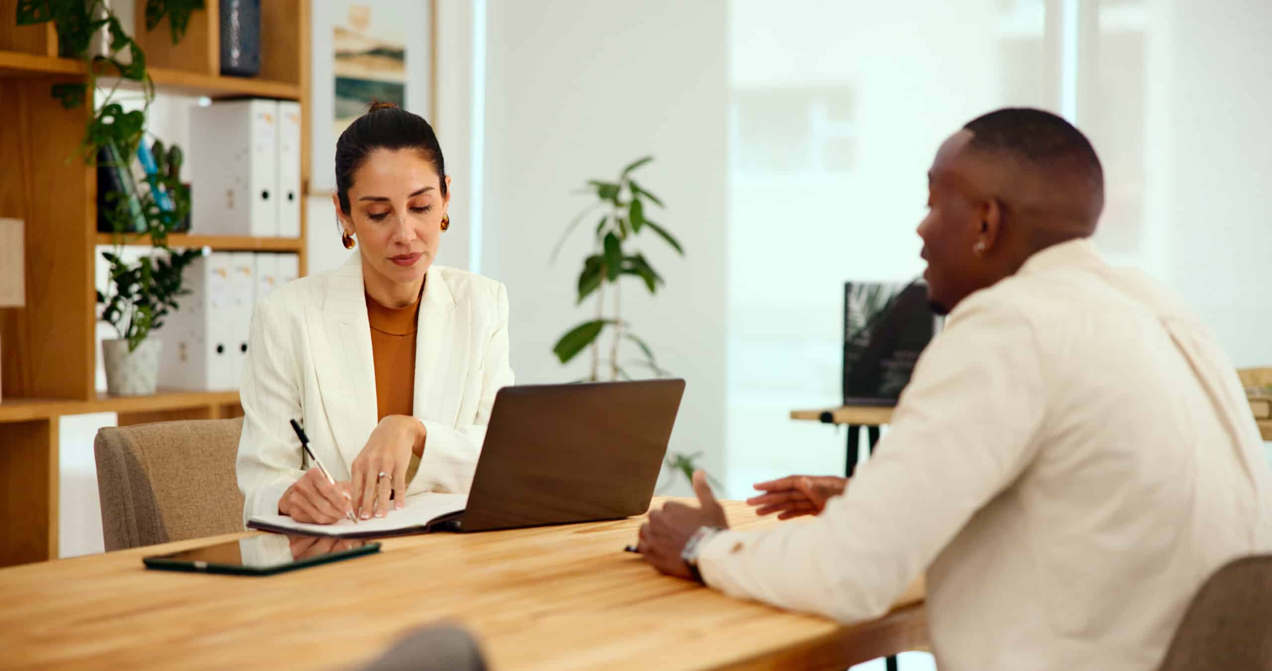 Femme en blazer blanc écrivant sur un carnet à côté d'un ordinateur portable, en réunion avec un homme, bureau lumineux