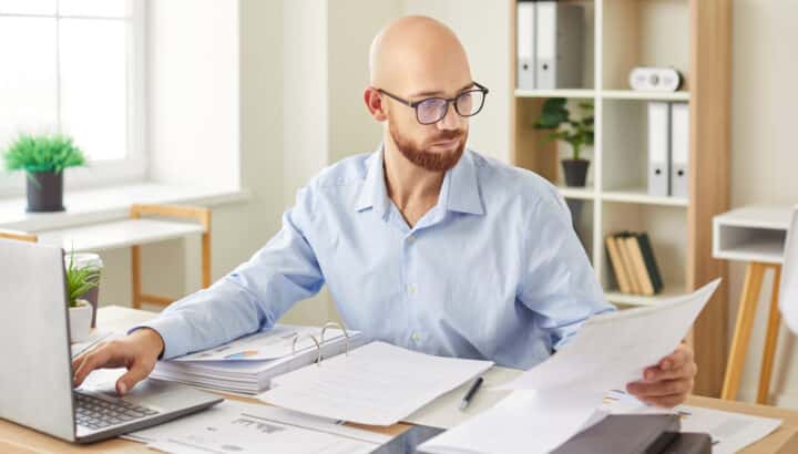 Homme chauve avec lunettes et barbe, consultant des documents papier et un classeur à anneaux, tapant sur un ordinateur portable