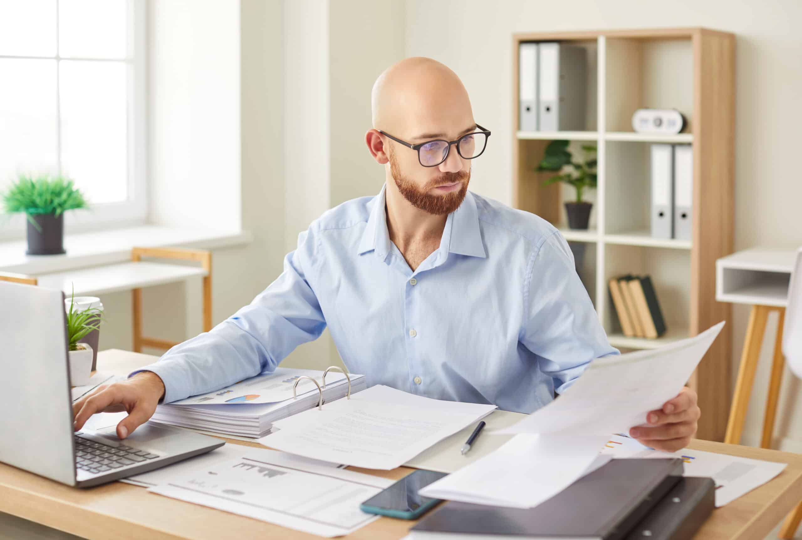 Homme chauve avec lunettes et barbe, consultant des documents papier et un classeur à anneaux, tapant sur un ordinateur portable